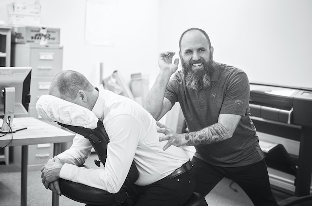 A smiling man performs massage to a clothed man in a massage chair in an office setting.