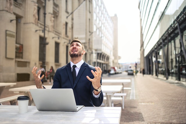 A man in a business suit is seated at a patio table between two tall buildings with his laptop and coffee cup on the table. He looks skyward with his hands extended in front of him, slightly curled, expressing a look of frustration and distress.