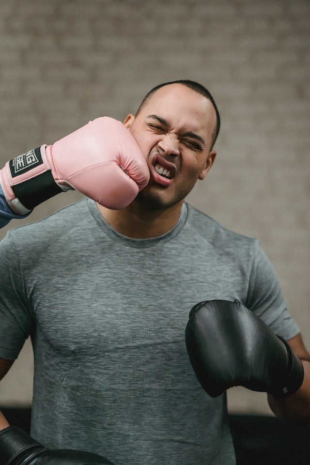 A man wearing boxing gloves makes a pained face on receiving a hit to the right cheek from a boxing glove worn by an unseen person out of frame.